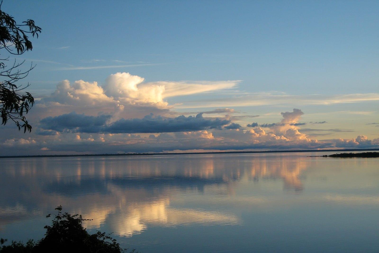 Sunset over a broad Amazon river, clouds mirrored in still water.